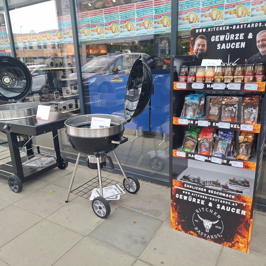Ein offener Grill steht vor einem Geschäft mit einem Schild, das Gewürze und Saucen bewirbt. Im Hintergrund ist ein Regal mit verschiedenen Produkten zu sehen.