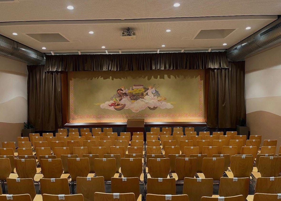 Ein leerer Theatersaal mit einer großen Bühnenkulisse, die ein Gemälde von einer Person in einem Boot auf einer Wolke zeigt. Die Stühle sind nummeriert und in Reihen angeordnet.