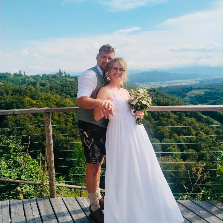 Ein frisch verheiratetes Paar steht auf einer Holzterrasse und blickt auf ein üppiges grünes Tal. Der Bräutigam trägt eine Weste und Shorts, während die Braut einen Blumenstrauß hält.