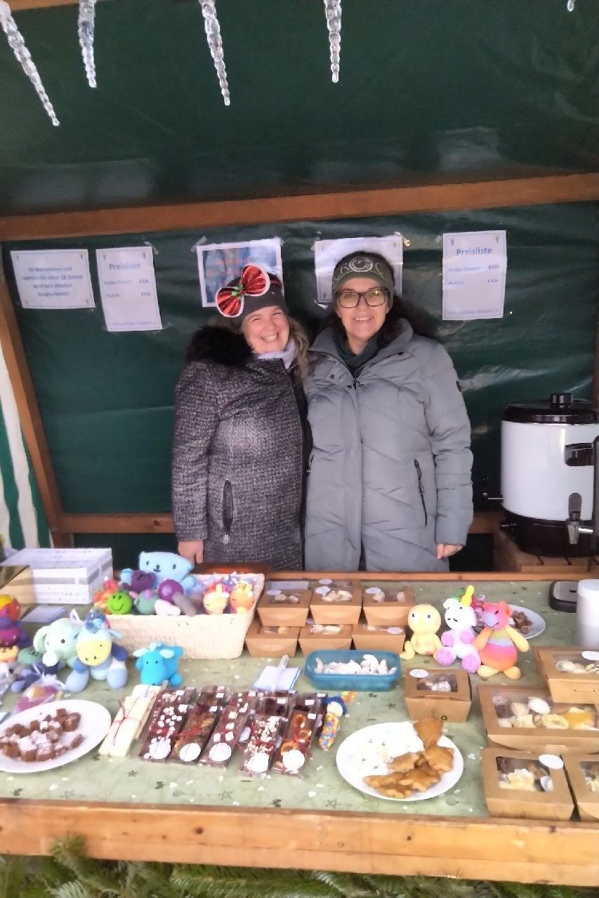 Two women stand in front of a stall with various items on display, including toys, chocolates, and cookies. They are both wearing winter jackets. Behind them is a green backdrop with price tags and posters.