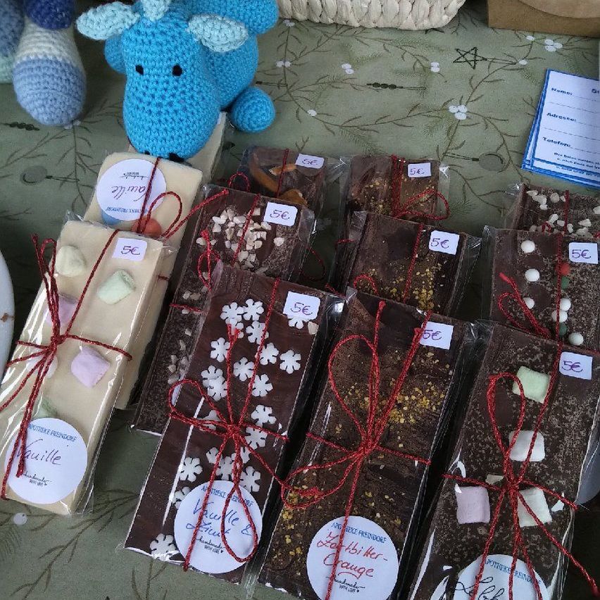 Various chocolate bars, wrapped with red ribbons and tied with red string, are on a table. Some have marshmallows, snowflakes, and other decorations. A blue stuffed animal and knitted items are also present.
