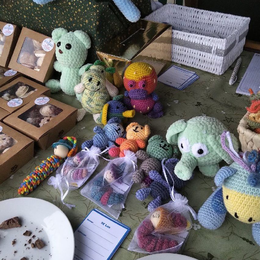 A table is filled with various crocheted toys, including bears, dragons, and other creatures. Some toys are in small plastic bags, while others are in boxes. A white plate with crumbs sits on the left.