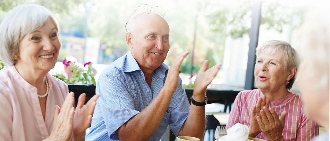 Drei ältere Menschen, zwei Frauen und ein Mann, sitzen zusammen und scheinen zu klatschen. Das Bild hat einen deutschen Textüberlagerung, der 'Wir freuen uns auf Euer Kommen!' mit einer Website-URL besagt.