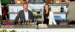 Ein Mann und eine Frau stehen hinter einer Theke in einem Weingut. Sie halten Weingläser. Regale mit Flaschen stehen dahinter. Ein Banner mit dem Text Raiding - Mittelburgenland ist davor.