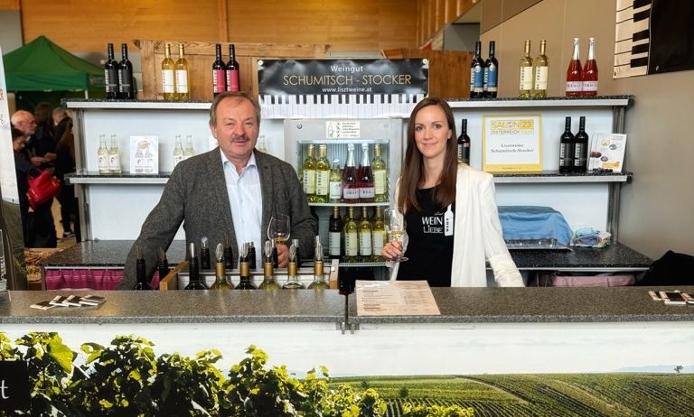 Ein Mann und eine Frau stehen hinter einer Theke in einem Weingut. Sie halten Weingläser. Regale mit Flaschen stehen dahinter. Ein Banner mit dem Text Raiding - Mittelburgenland ist davor.