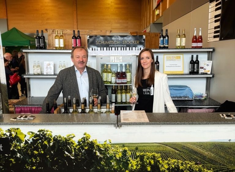 Ein Mann und eine Frau stehen hinter einer Theke in einem Weingut. Sie halten Weingläser. Regale mit Flaschen stehen dahinter. Ein Banner mit dem Text Raiding - Mittelburgenland ist davor.