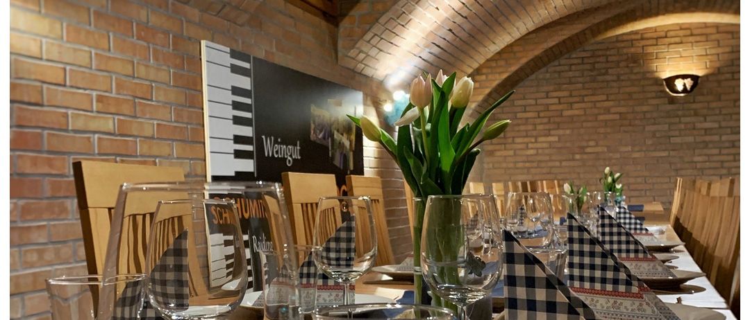 Ein Weinkeller für Sie, eine Veranstaltung im privaten Rahmen. Ein Tisch mit Weingläsern, Servietten und Tellern. Eine Ziegelwand mit Poster und Lampen.