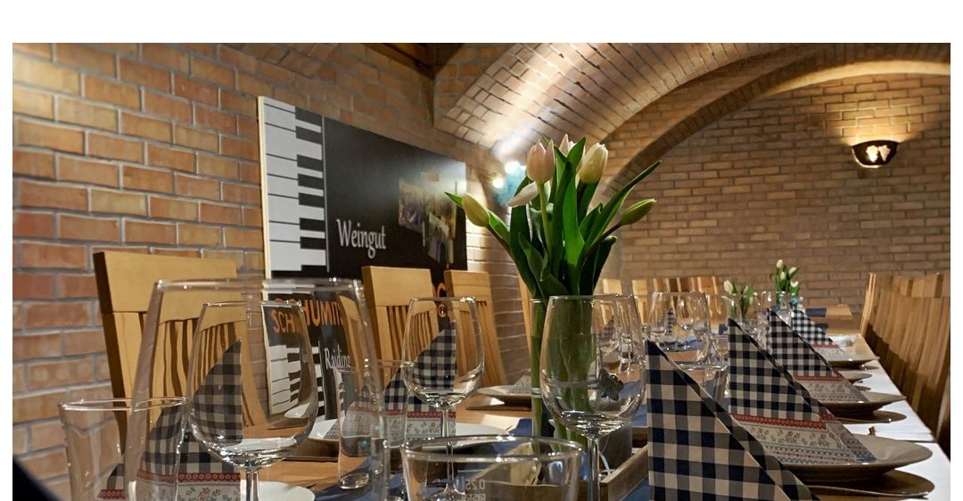 Ein Weinkeller für Sie, eine Veranstaltung im privaten Rahmen. Ein Tisch mit Weingläsern, Servietten und Tellern. Eine Ziegelwand mit Poster und Lampen.