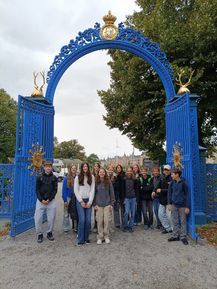Eine Gruppe von Schülern steht vor einem blauen Tor mit goldenen Hirschköpfen oben. Sie sind lässig gekleidet und scheinen für ein Foto zu posieren.
