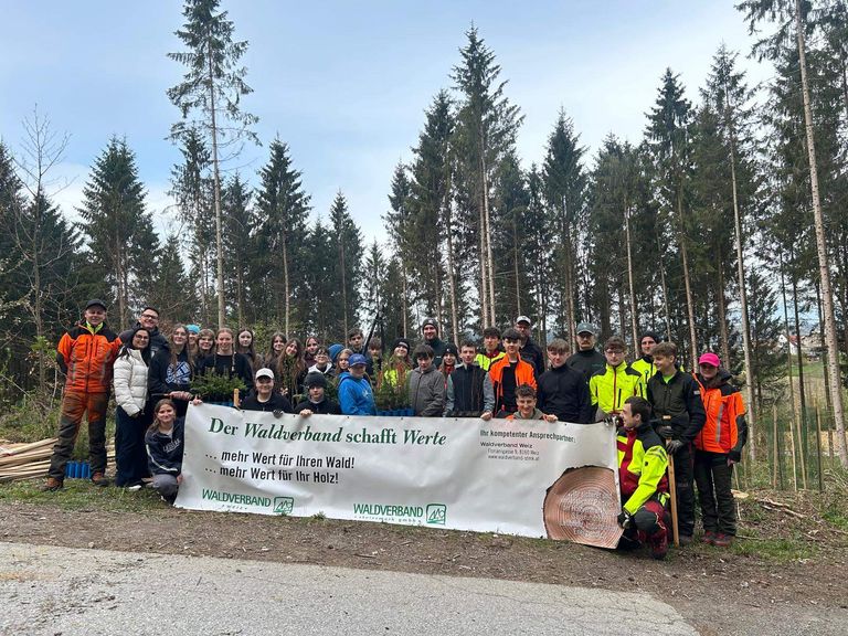 Eine Gruppe von Menschen in Schutzausrüstung posiert für ein Foto vor einem Wald mit einem Banner, das 'Der Waldverband schafft Werte' steht. Das Banner wird von zwei Personen in der Mitte gehalten.