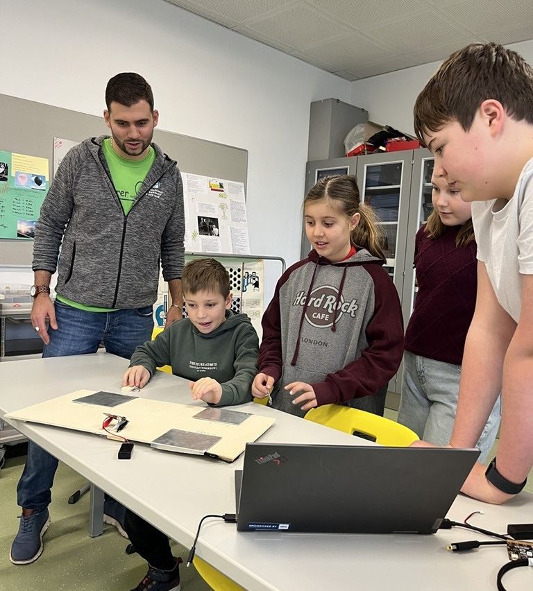 Four children and a man are gathered around a table in a classroom. They are looking at a laptop and a solar panel. The children are wearing jackets.