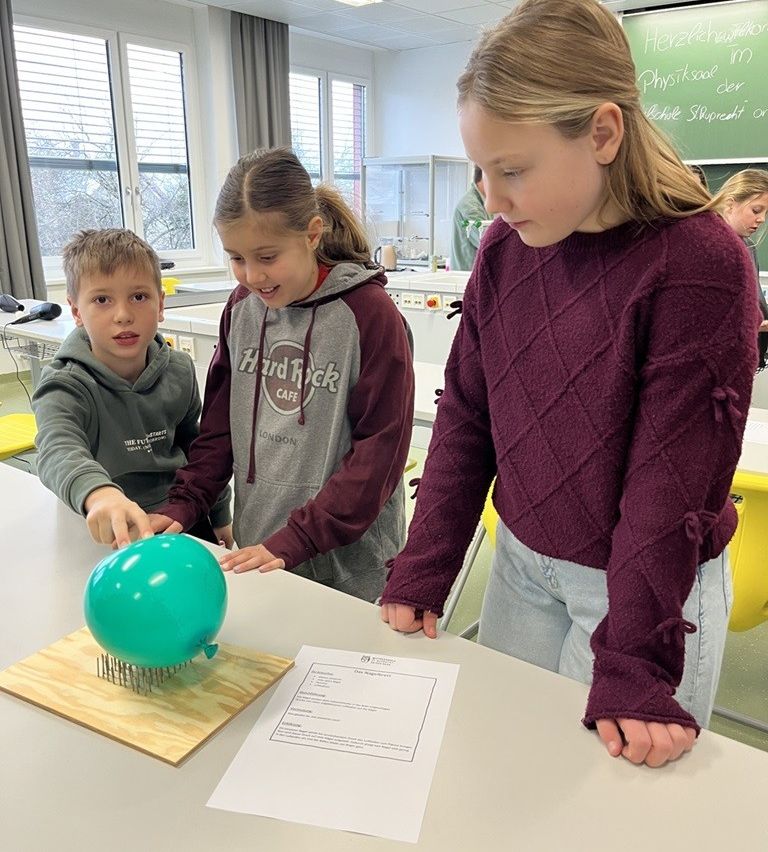 Three children are in a classroom. A green balloon is on a board with nails. The kids are looking at it.