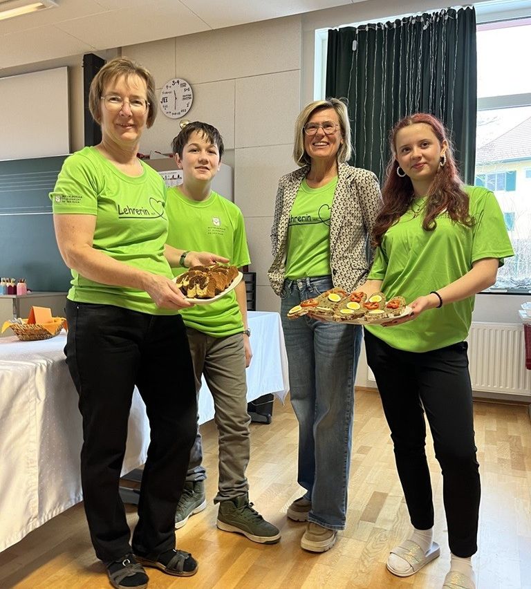 Three women and a boy in green shirts holding plates of food stand in a room with a clock and window.