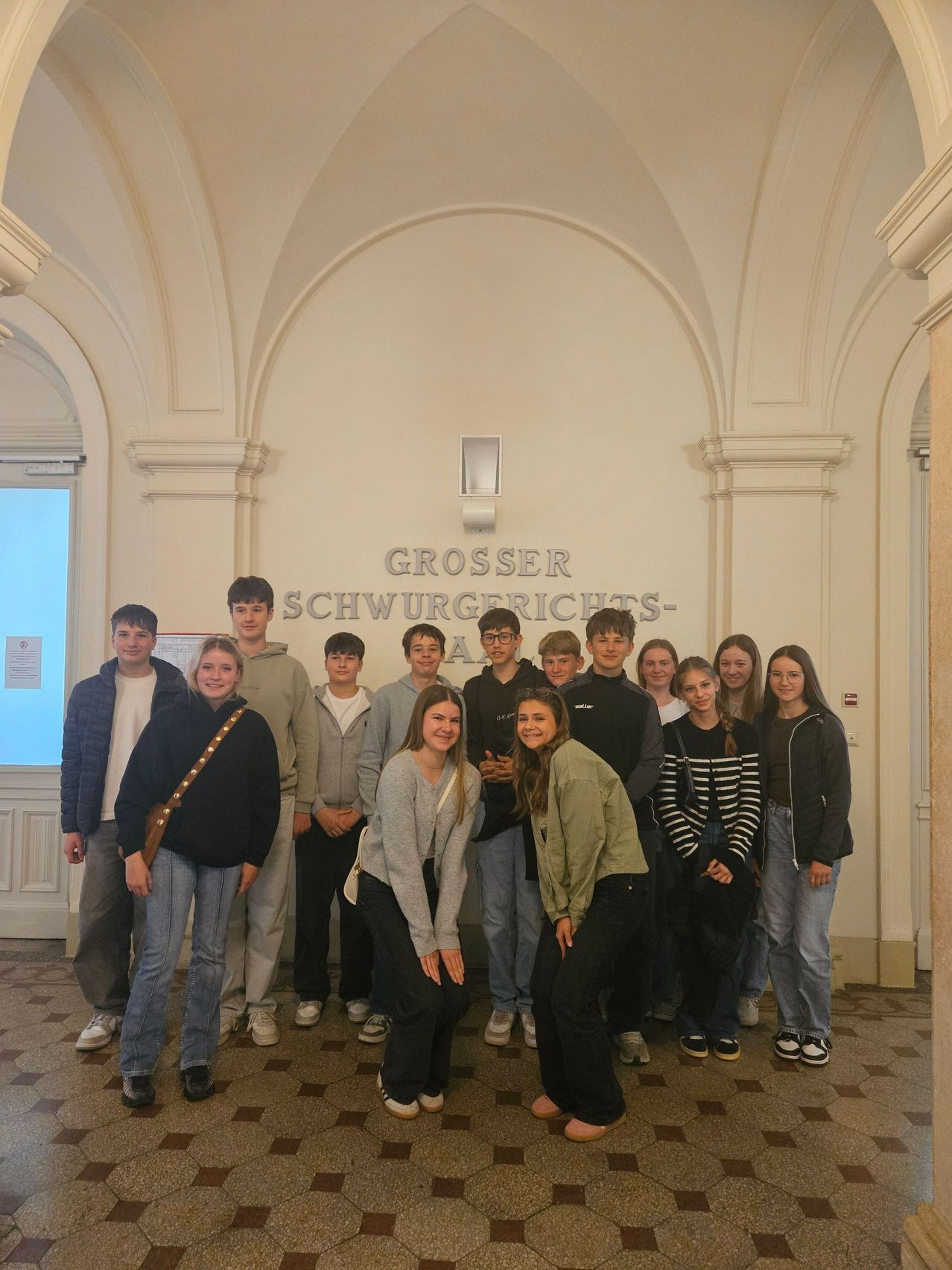 A group of young people are posing for a photo in front of a wall with the text 'Grosser Schwurgerichts-A'. They are wearing casual clothes and smiling.