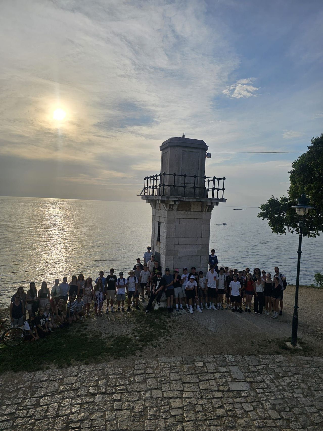 A group of people stands in front of a lighthouse on a sunny day, with the ocean in the background.