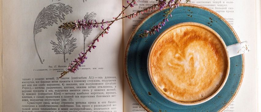 Ein offenes Buch mit botanischen Illustrationen liegt flach, mit einer Tasse Kaffee auf einer blauen Untertasse darauf. Der Kaffee hat Milchkunst, und lila Blumen sind auf der Untertasse.