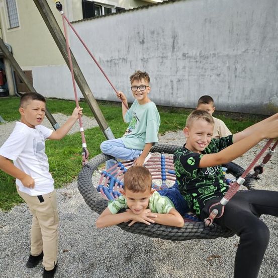 Fünf Jungen spielen auf einer Reifenschaukel im Spielplatz, einer von ihnen lächelt. Ein anderer Junge steht neben der Schaukel.
