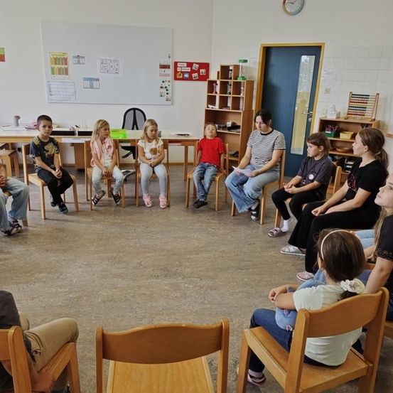 Eine Gruppe von Kindern und eine Lehrerin sitzen in einem Klassenzimmer im Kreis. Die Lehrerin hält ein Papier in der Hand und die Kinder hören aufmerksam zu. Der Raum hat Holzstühle, eine Tafel und Regale.