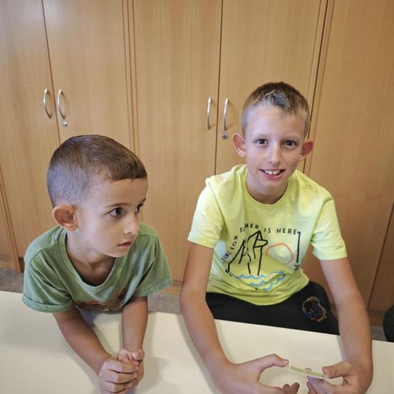Zwei Jungen, einer mit kurzen Haaren in einem grünen T-Shirt und der andere in einem gelben T-Shirt, sitzen an einem Tisch, möglicherweise spielen sie ein Spiel.