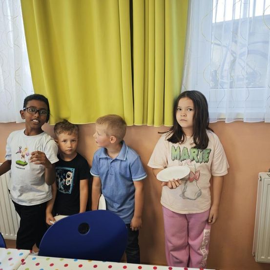 Vier Kinder stehen nebeneinander, zwei halten Teller. Ein Mädchen hält einen Teller mit einem Hasenbild. Der Junge trägt eine Brille. Sie stehen vor einer Wand mit einem Fenster.