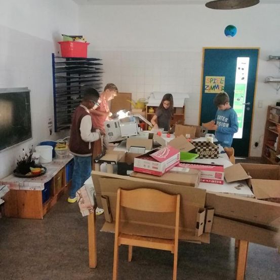 Vier Kinder arbeiten in einem Raum und ordnen Gegenstände auf einem Tisch mit Kisten und Bastelmaterial an. Der Raum hat Regale, eine Tür und eine Tafel.