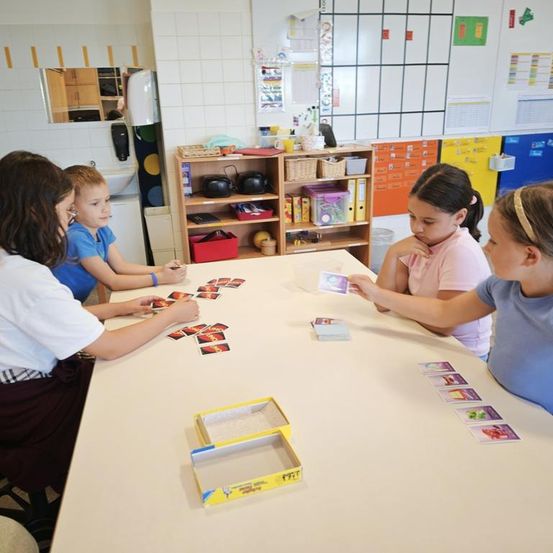 Vier Kinder spielen ein Kartenspiel auf einem Tisch in einem Klassenzimmer, mit einer Schachtel und Karten auf dem Tisch und einem Regal mit Gegenständen dahinter.