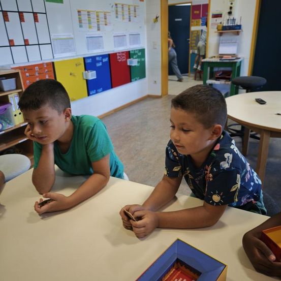 Zwei Jungen sitzen an einem Tisch und spielen ein Kartenspiel in einem Klassenzimmer mit farbenfrohen Wandschränken und einer Person, die im Hintergrund geht.