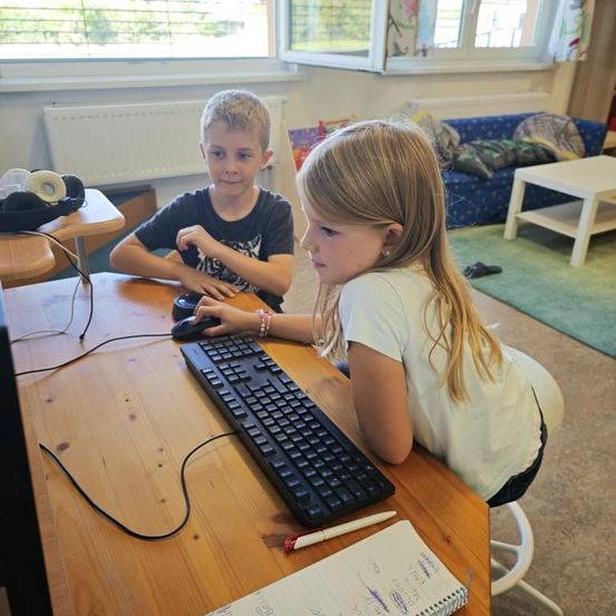 Zwei Kinder sitzen an einem Schreibtisch und benutzen einen Computer, ein Mädchen tippt auf der Tastatur, ein Junge lächelt neben ihr, Kopfhörer auf dem Tisch und ein Notizbuch vor ihnen.