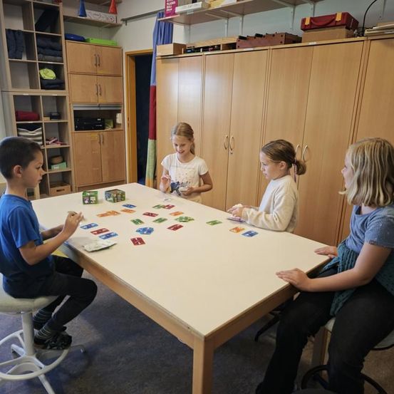 Vier Kinder sitzen um einen Tisch herum und spielen ein Kartenspiel, Karten verstreut auf dem Tisch. Der Raum hat Holzschränke und Regale.