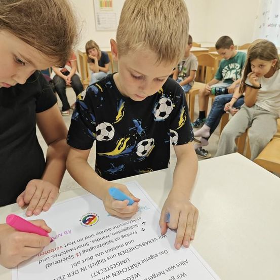 Kinder schreiben auf einem großen Stück Papier auf einem Tisch in einem Klassenzimmer. Ein Kind hält einen rosa Marker.