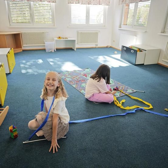 Zwei junge Mädchen spielen mit Bändern auf einem Teppich in einem Raum mit weißen Wänden und Heizkörpern.