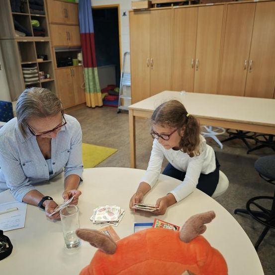 Zwei Personen, eine Frau und ein Mädchen, sitzen um einen Tisch herum in einer Innenumgebung. Die Frau trägt eine Brille und eine Uhr, während das Mädchen eine Brille trägt und eine Karte in der Hand hält. Ein Plüschtier liegt auf dem Tisch. Hinter ihnen steht ein Holzschrank mit Regalen.