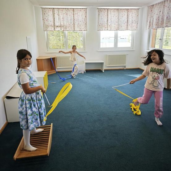 Drei Mädchen spielen mit gelben Bändern in einem Raum mit blauem Teppich und weißen Wänden. Ein Mädchen steht auf einer Holzplattform.