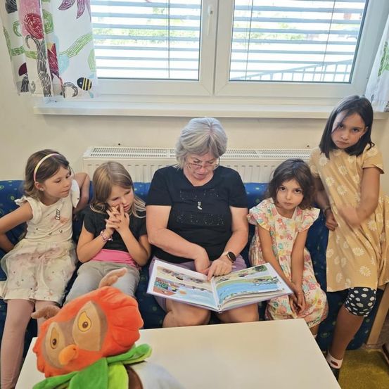 Eine ältere Frau liest einem kleinen Mädchen vor, das auf einem blauen Sofa mit einer Gruppe junger Mädchen sitzt. Ein Kuscheltier steht auf einem Tisch vor ihnen. Das Zimmer hat ein Fenster mit Jalousien.