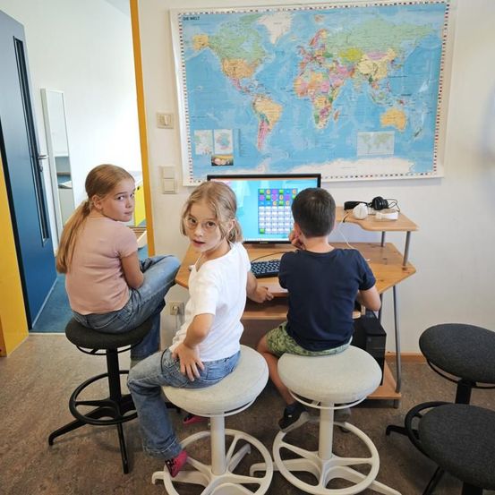 Drei Kinder sitzen auf Hockern in einem Raum. Eines benutzt einen Computer, während die anderen beiden zusehen. Eine Weltkarte hängt an der Wand.