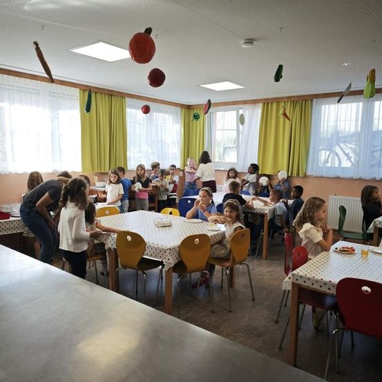 Ein Klassenzimmer ist mit Kindern gefüllt, die an Tischen sitzen, einige mit Tellern und andere mit Papiertellern. Bunte Dekorationen hängen von der Decke.