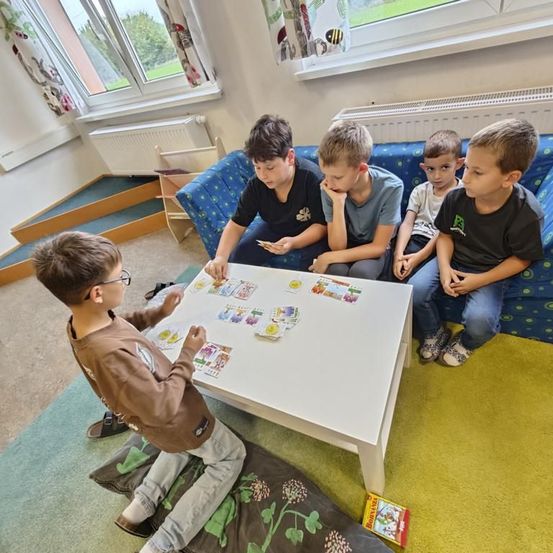 Fünf Jungen sitzen auf einer blauen Couch und dem Boden und spielen ein Kartenspiel auf einem weißen Tisch. Ein Junge trägt eine Brille.