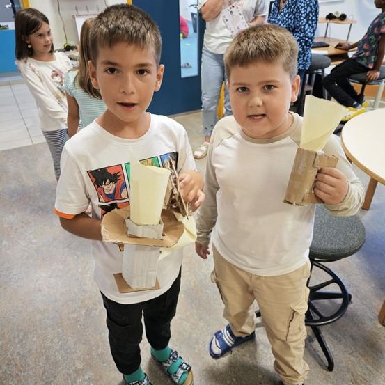 Zwei Jungen in einem Klassenzimmer halten Papierbasteleien hoch, eine mit einem Kegel und die andere mit einem Becher. Hinter ihnen arbeiten andere Kinder an Tischen.