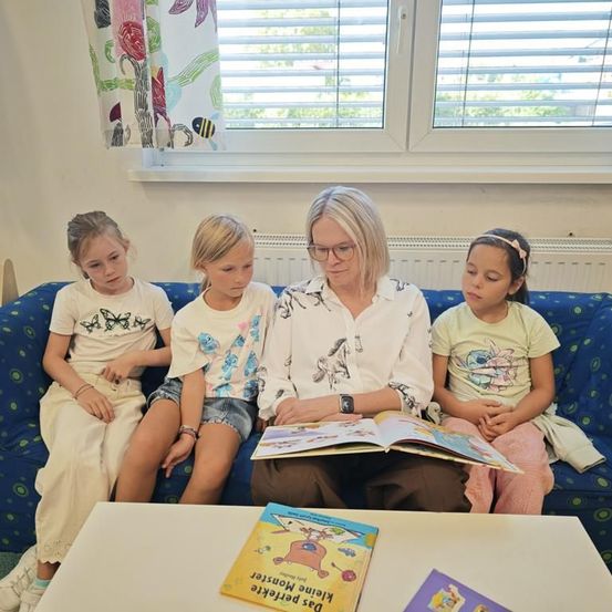 Eine erwachsene Frau liest drei Mädchen auf einer Couch in einem Raum ein Buch vor. Die Frau trägt eine Brille und eine Uhr. Die Mädchen tragen weiße und gelbe Hemden. Ein Buch liegt auf dem Tisch.