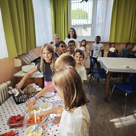 Eine Gruppe von Kindern versammelt sich um einen Tisch mit verschiedenen Lebensmitteln. Einige Kinder stehen, während andere am Tisch sitzen. Ein Kind hält einen Tablett mit Essen.