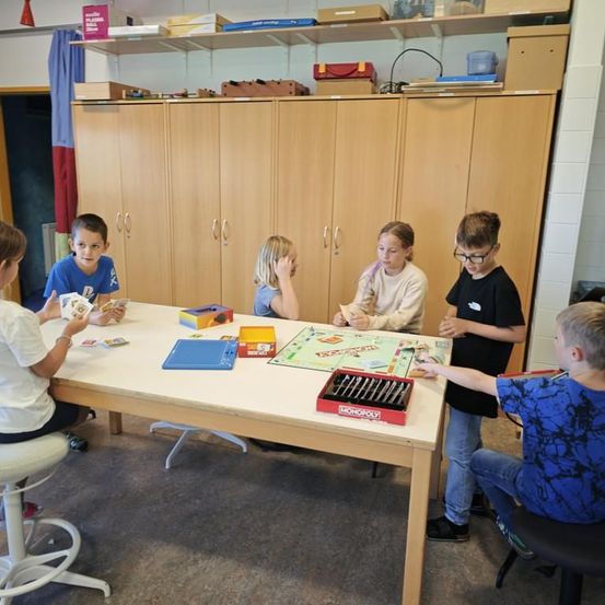 Eine Gruppe von Kindern spielt ein Brettspiel in einem Raum. Sie sitzen um einen Tisch mit Monopoly und anderen Spielzeugen. Der Raum hat Holzschränke mit Regalen darüber.