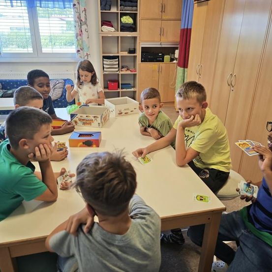 Eine Gruppe von Kindern sitzt um einen Tisch herum und spielt ein Kartenspiel, mit Schachteln und Karten auf dem Tisch. Der Raum hat Holzschränke und Regale.