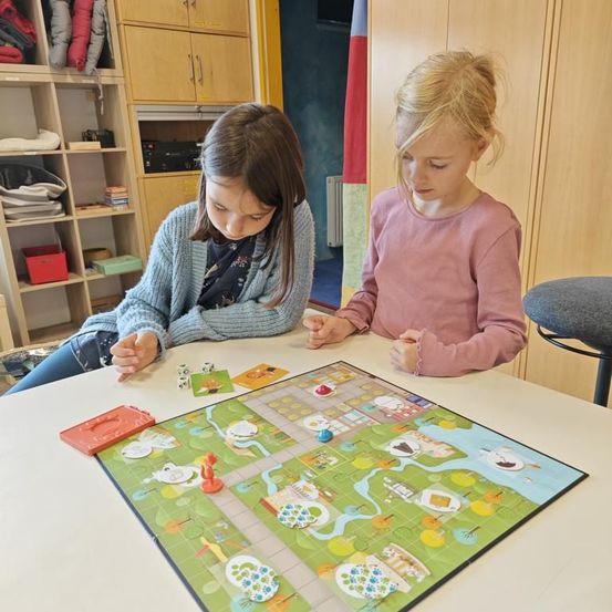 Zwei junge Mädchen sitzen an einem Tisch und spielen ein Brettspiel. Das Mädchen auf der linken Seite konzentriert sich auf das Spiel, während das Mädchen auf der rechten Seite neugierig aussieht. Der Tisch hat verschiedene Spielsteine. Der Hintergrund zeigt einen Raum mit Holzschränken und Regalen.