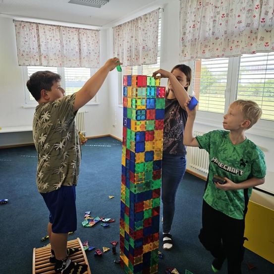 Drei Kinder stehen um eine große, bunte würfelförmige Struktur herum, möglicherweise bauen sie sie. Ein Kind auf der linken Seite hält ein grünes Objekt. Ein weiteres Kind in der Mitte hält ein blaues Objekt. Das dritte Kind auf der rechten Seite hält ein Minecraft-Objekt.