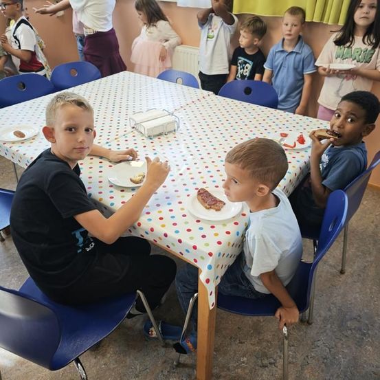 Mehrere Kinder sitzen am Tisch und essen. Ein Junge in einem schwarzen T-Shirt schaut in die Kamera. Ein anderer Junge isst Kuchen, während ein Mädchen zusieht.