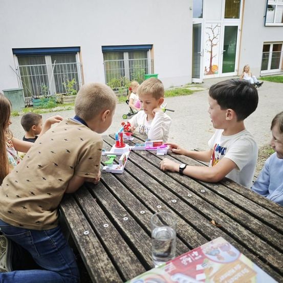 Mehrere Kinder spielen ein Brettspiel auf einem Holztisch in einem Außenbereich. Ein junges Mädchen sitzt in der Nähe eines Kinderwagens. Ein Gebäude mit Glastüren und Fenstern befindet sich dahinter.
