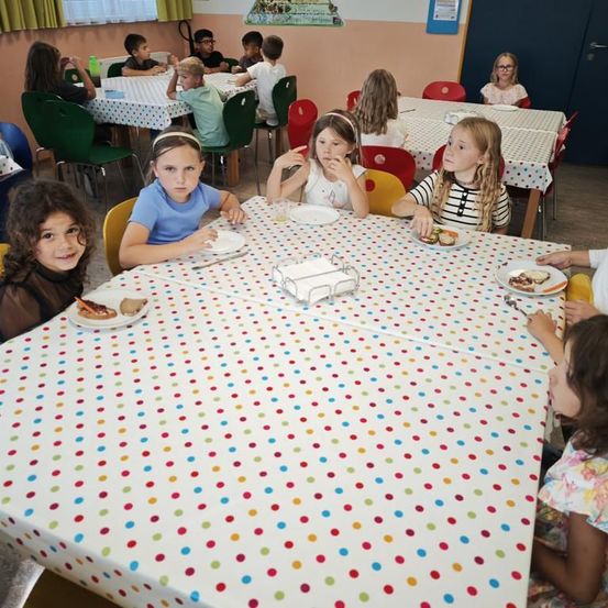 Ein Klassenzimmer mit Schülern, die an Tischen sitzen und essen. Die Tische sind mit farbenfrohen Tupfentischdecken bedeckt. Einige Schüler tragen eine Brille. Das Klassenzimmer hat Glasfenster und eine Tür.
