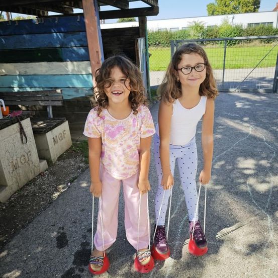 Zwei junge Mädchen mit Brille lächeln und stehen auf roten Skiern im Freien. Sie halten die Griffe der Skier. Hinter ihnen steht eine Holzbank und eine Holzkonstruktion mit der Aufschrift 'MGF'.