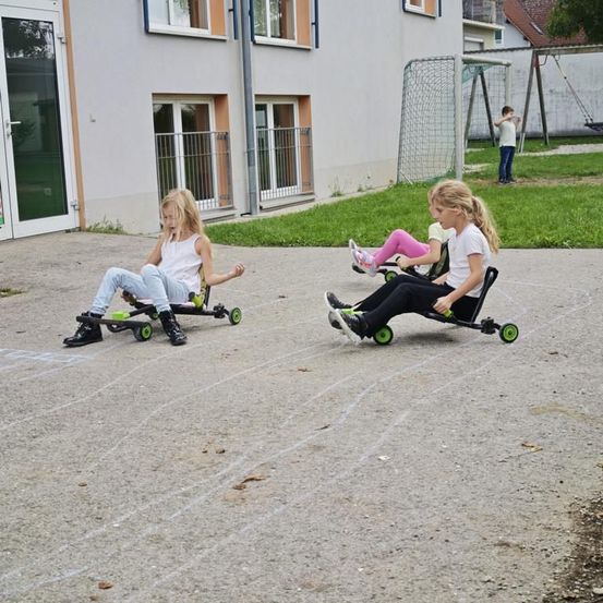 Drei Kinder fahren Go-Karts auf einer Einfahrt vor einem Gebäude mit Fenstern und einem Rasenbereich. Ein Kind steht in der Nähe eines Tors und schaukelt.