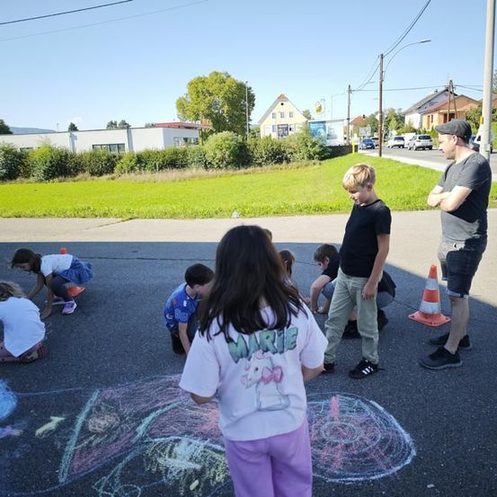 Mehrere Kinder zeichnen mit Kreide auf der Straße, während zwei Erwachsene in der Nähe stehen. Im Hintergrund sind eine Wiese und Häuser zu sehen.